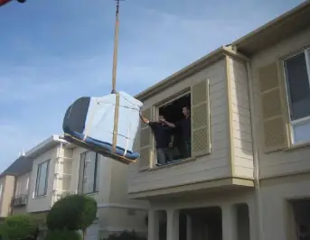 A large, wrapped item is being hoisted out of an upper window by a crane, while two people assist from inside the house