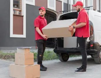 Two delivery workers in red shirts are transferring a large cardboard box from a van to the ground, with stacked boxes nearby